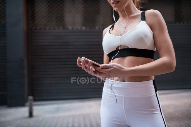 Muscular woman in white sportive outfit using earphones and smartphone on street — Stock Photo
