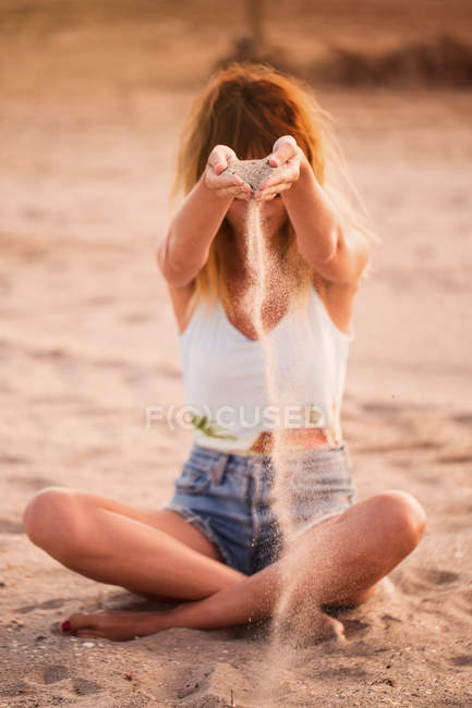 Donna in pantaloncini di jeans seduta sulla spiaggia sabbiosa e versando mucchio di sabbia attraverso le mani — Foto stock