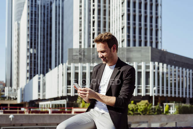 Elegante chico sonriente navegando teléfono inteligente en la calle de la ciudad moderna - foto de stock