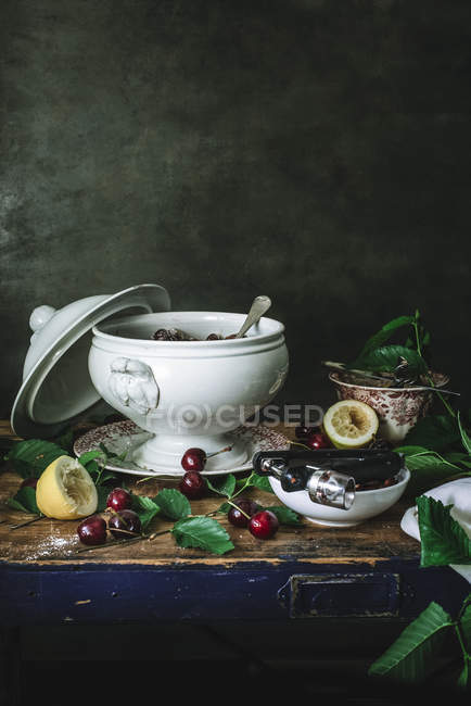 Composition of ceramic bowl with cherries and sugar and cherry kernel remover on table with green leaves — Stock Photo