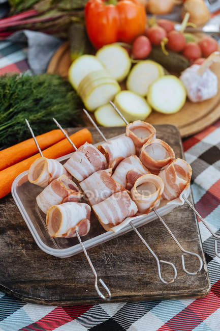 Close-up of metal skewers with strips of bacon on dish on wooden board — Stock Photo