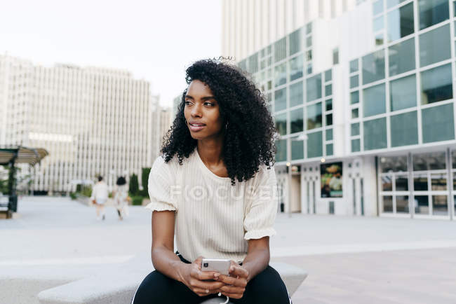 Thoughtful African-American woman sitting on city street and holding smartphone — Stock Photo