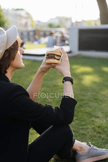 Mulher elegante sentado na grama do parque e segurando delicioso hambúrguer takeaway — Fotografia de Stock