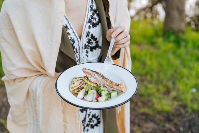 Frau hält Teller mit frischem Gemüse mit gegrilltem Lachsfilet und Zucchini auf Picknick — Stockfoto