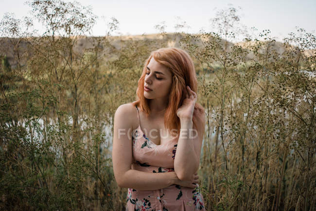 Рыжая молодая женщина в декоративном платье трогает волосы и позирует на природе — стоковое фото