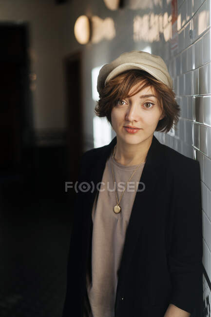 Junge Frau mit Kurzhaarfrisur in schlichter schwarzer Jacke und beiger Mütze blickt in die Kamera und lehnt an der Wand — Stockfoto