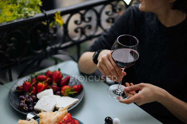 Vista de colheita de fêmea em blusa listrada segurando elegante copo de vinho branco sentado à mesa de tábua de madeira com bebida brilhando no fundo borrado — Fotografia de Stock
