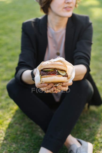 Mulher segurando hambúrguer enquanto sentado na grama — Fotografia de Stock