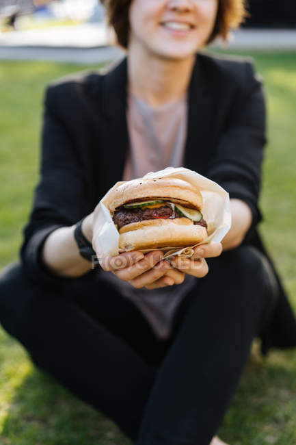 Lächelnde Frau zeigt Burger, während sie im Park auf Gras sitzt — Stockfoto
