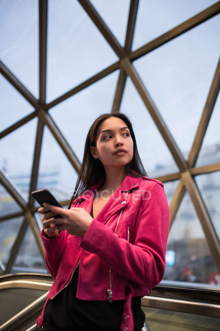 Красивая молодая женщина в розовой куртке стоит со смартфоном в современном здании и смотрит в сторону — стоковое фото