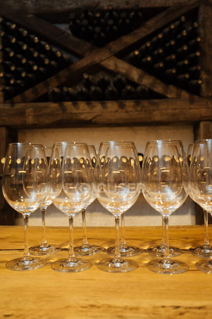 Рядок елегантних блискучих окулярів, що стоять на дерев'яному столі у винному підвалі з пляшками вина на полицях на фоні — стокове фото
