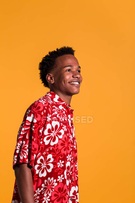 Sonriente joven afroamericano hombre vistiendo camisa de playa colorida y mirando hacia otro lado sobre fondo naranja - foto de stock