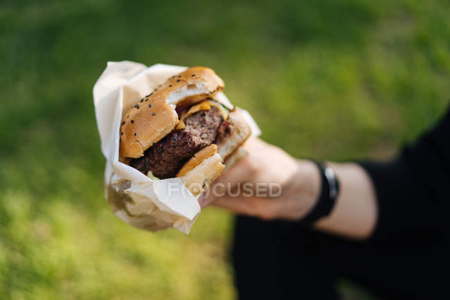 Main humaine tenant hamburger tout en étant assis sur l'herbe — Photo de stock