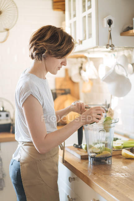 Donna mettendo ingredienti in tazza di plastica del frullatore per fare frullato verde sano — Foto stock