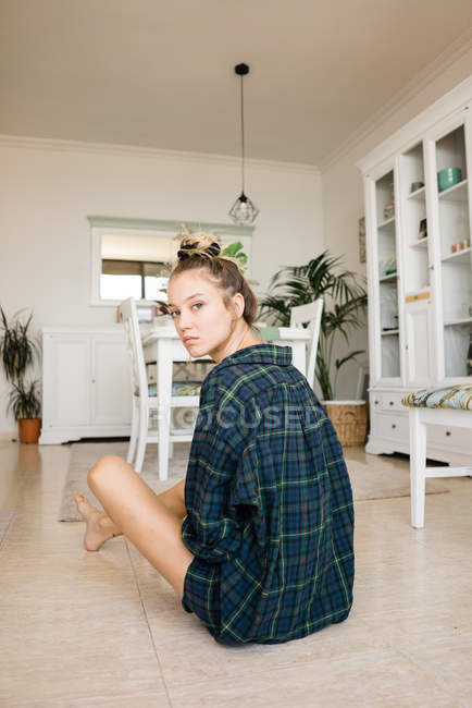 Mujer joven con camisa a cuadros sentada en el suelo en una habitación elegante y mirando a la cámara - foto de stock