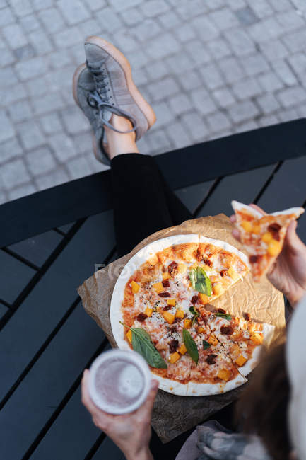 Mujer sentada en el podio en la calle con un vaso de cerveza y pizza para llevar - foto de stock