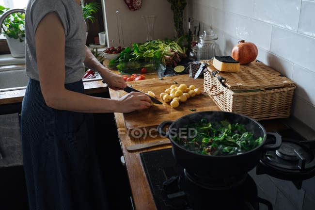 Donna che taglia patate sul tagliere di legno e foglie di spinaci in pentola sul fornello a gas — Foto stock