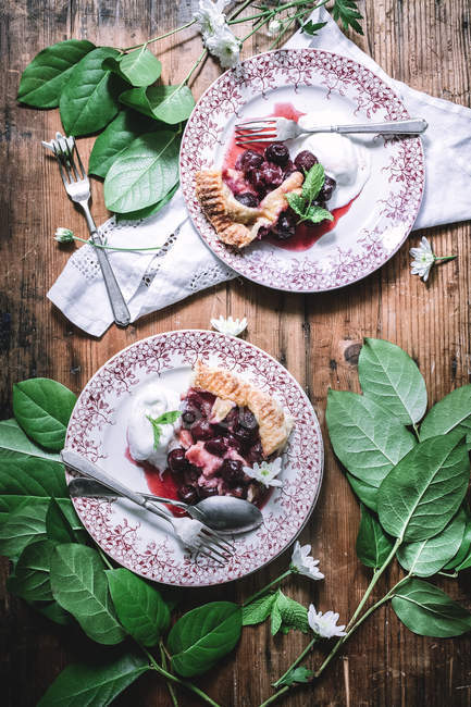 Top view of plates with cherry pie slices served with ice cream on table among green leaves — Stock Photo
