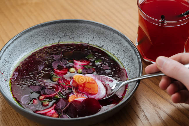 Human hand holding metal spoon over plate of Nordic beetroot soup in grey bowl on wooden table with drink — Stock Photo