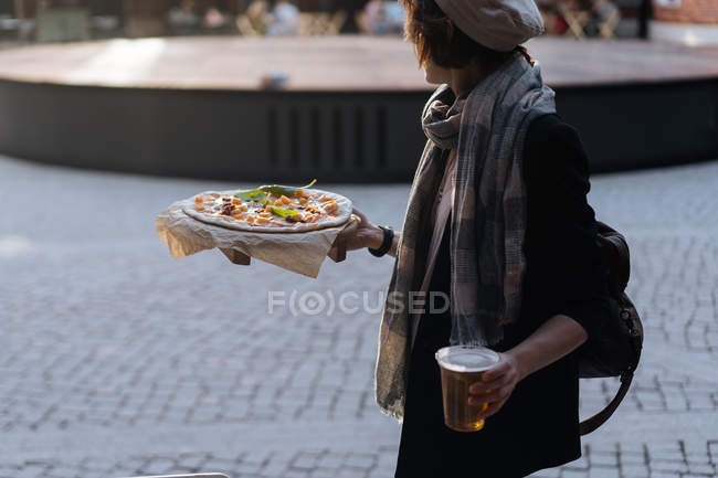 Woman holding glass of beer and pizza while walking in outside cafeteria — Stock Photo
