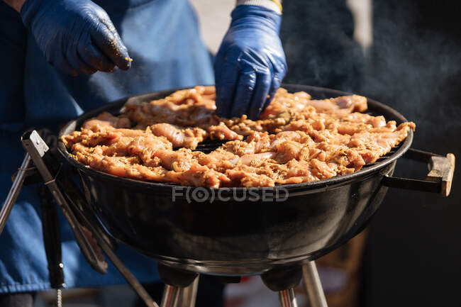 Faceless shot of man in gloves arraying raw meat on metal grid in barbecue grill standing in sunlight — Stock Photo