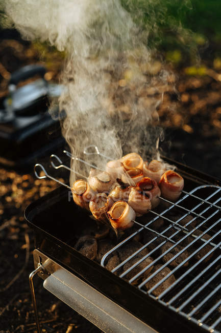 Nahaufnahme einer tragbaren Grillpfanne mit brennender Holzkohle und Spießen mit Speckstreifen beim Grillen — Stockfoto