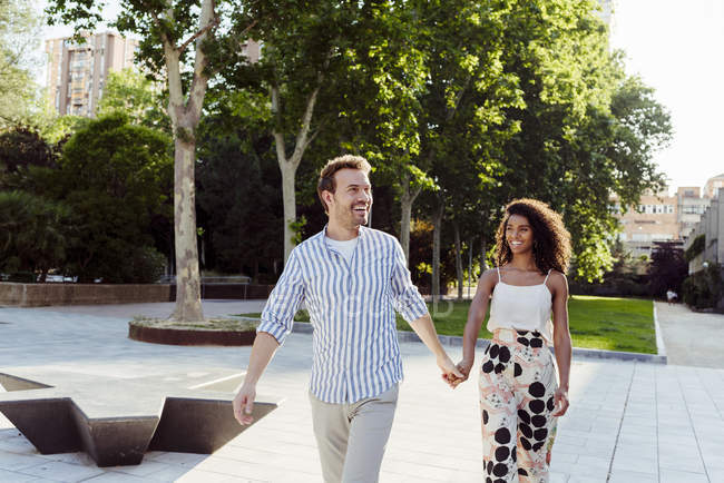 Sonriente pareja multirracial caminando de la mano en el parque de la ciudad - foto de stock