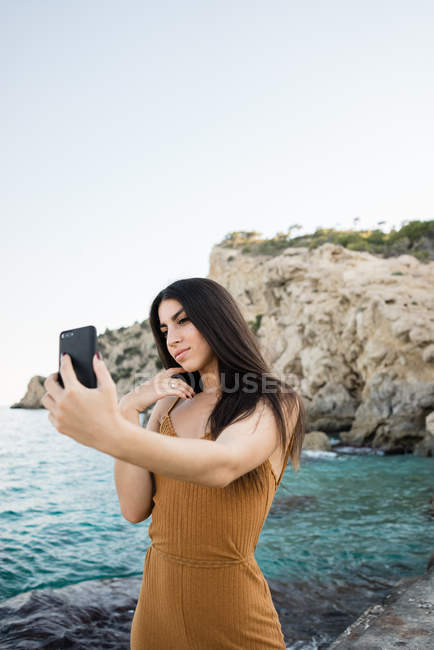 Stylish young brunette taking selfie on beach by sea water — Stock Photo