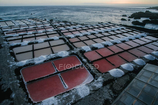 Vista pitoresca de pequenas lagoas de cristalização retangulares com água do oceano e sal produzido em pilhas ao redor na costa do mar rochoso ao pôr do sol em Espanha — Fotografia de Stock