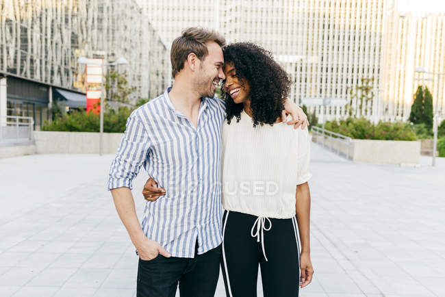 Alegre pareja multirracial abrazando y riendo mientras caminan juntos por la calle de la ciudad - foto de stock