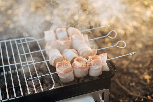 Primo piano della griglia portatile con carbone ardente e spiedini con strisce di pancetta grigliate — Foto stock