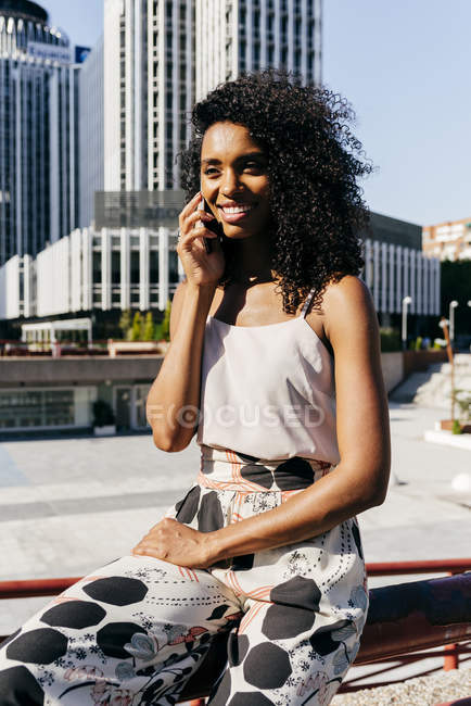 Elegant African-American woman smiling and talking on smartphone while sitting on fence on city street — Stock Photo