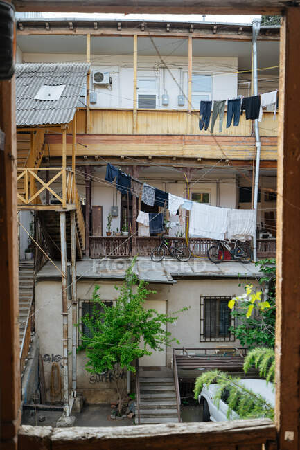 Вид через старое окно изношенного засохшего дома со свежепостроенным деревянным верхним этажом, изогнутыми лестницами и обветшалыми стенами с граффити, велосипедами, размещенными на террасе, и одеждой, высыхающей на стиральных линиях — стоковое фото