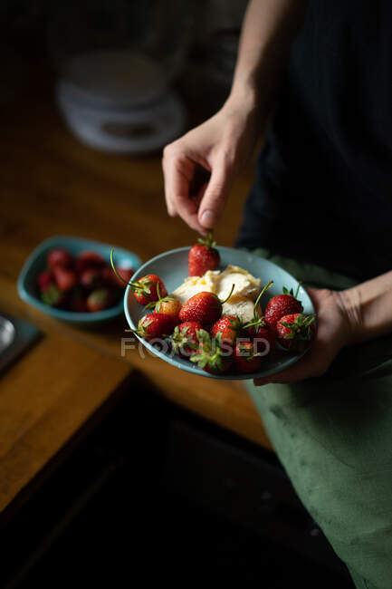 Vista de colheita de mão elegantemente tomando haste verde de morango suculento maduro de pequena tigela cheia de bagas vermelhas na mesa de madeira — Fotografia de Stock