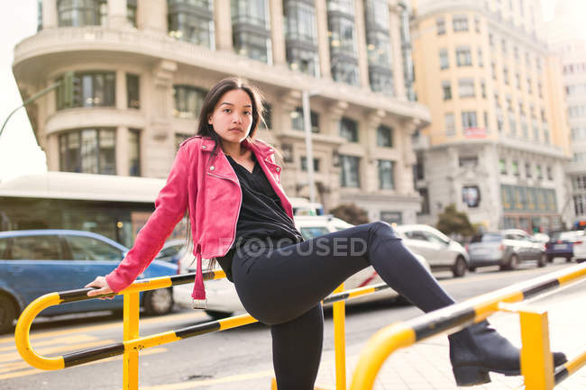 Молодая стильная женщина в черном и розовом пиджаке, опирающаяся на ограду и смотрящая в камеру — стоковое фото