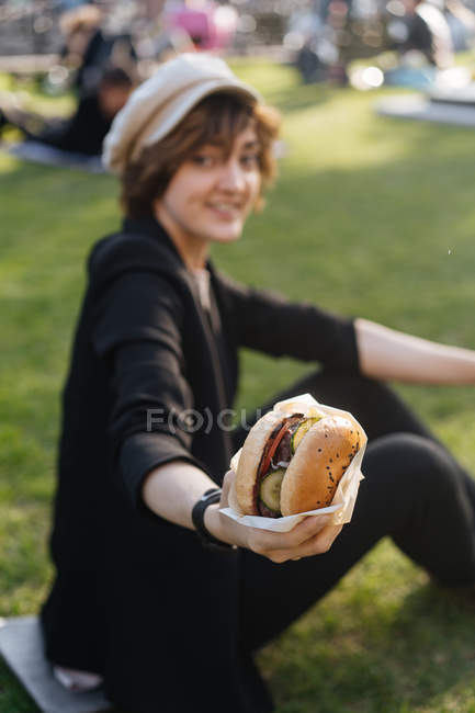 Femme souriante montrant hamburger tout en étant assis sur l'herbe dans le parc — Photo de stock