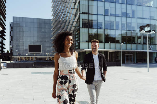 Sonriente elegante pareja multirracial cogido de la mano mientras camina en la ciudad moderna - foto de stock