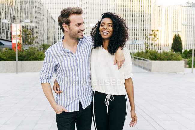 Cheerful multiracial couple hugging and laughing while walking on city street together — Stock Photo