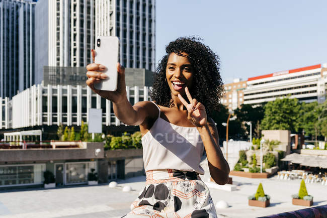 Mulher afro-americana elegante posando para selfie enquanto sentado na cerca na rua da cidade no dia ensolarado — Fotografia de Stock