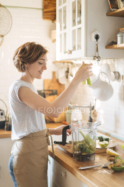 Femme mettant des ingrédients dans une tasse en plastique de mélangeur pour faire un smoothie vert sain — Photo de stock