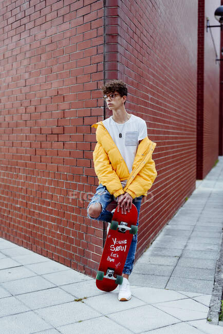 Молодий хлопчик-підліток в окулярах зі скейтбордом, що спирається на розі на вулиці — стокове фото