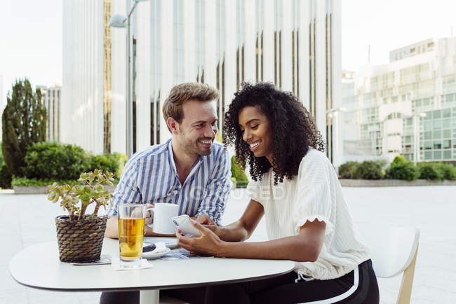 Nettes multiethnisches Paar lächelt und surft mit modernem Smartphone, während es gemeinsam am Tisch im Café sitzt — Stockfoto