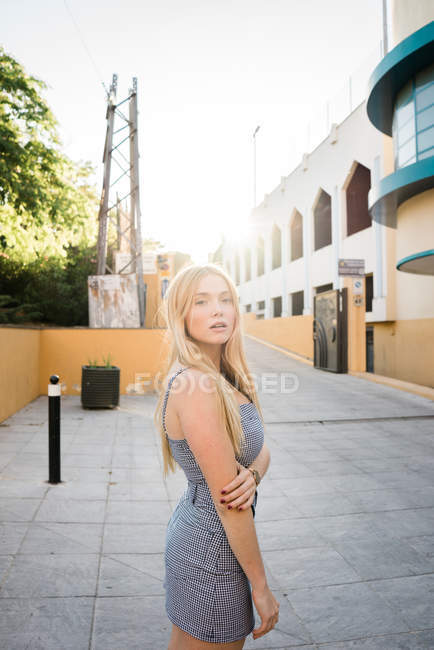 Blonde young woman in summer dress standing on street and looking at camera — Stock Photo