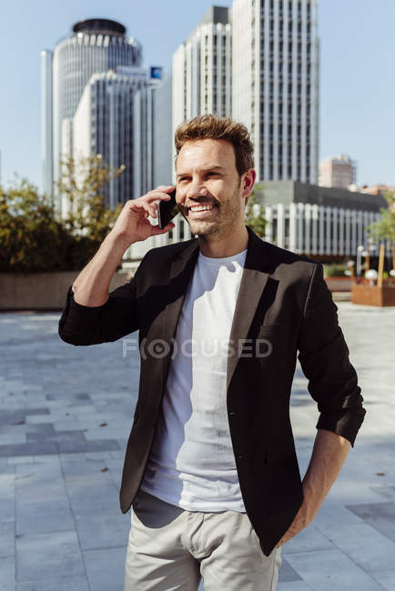 Lächelnder eleganter Mann, der am Telefon lächelt, während er an einem sonnigen Tag auf der Straße der modernen Stadt steht — Stockfoto