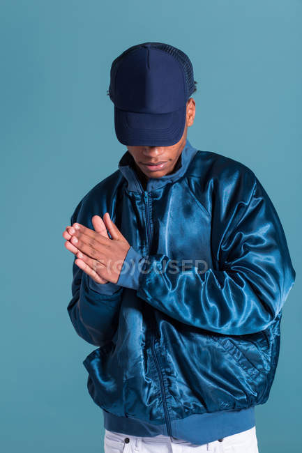 Trendiger ethnischer Mann mit glänzend blauer Bomberjacke und Mütze vor blauem Hintergrund — Stockfoto