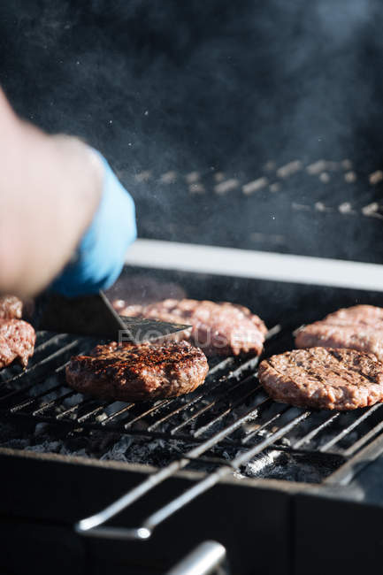 Human hand cooking raw burger patties roasting on grid of barbecue grill outdoors — Stock Photo