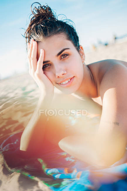 Sonriente chica joven tumbada en la playa a la luz del sol y mirando a la cámara - foto de stock
