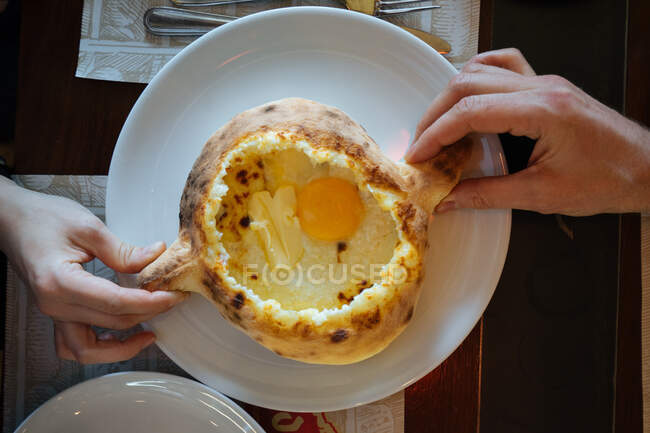 Crop view of male and female hands taking handles of traditional Georgian bread pot with fried eggs inside served on white ceramic plate on wooden table from above — Stock Photo