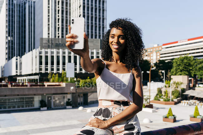 Elegant African-American woman posing for selfie while sitting on fence on city street on sunny day — Stock Photo