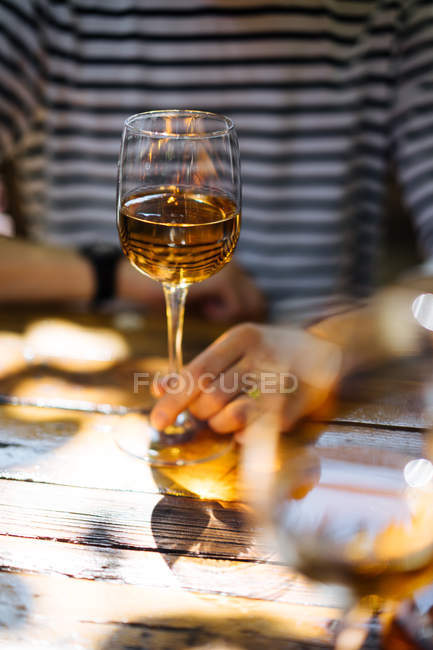 Mão feminina segurando vidro de vinho branco na mesa de madeira na luz solar ao ar livre — Fotografia de Stock